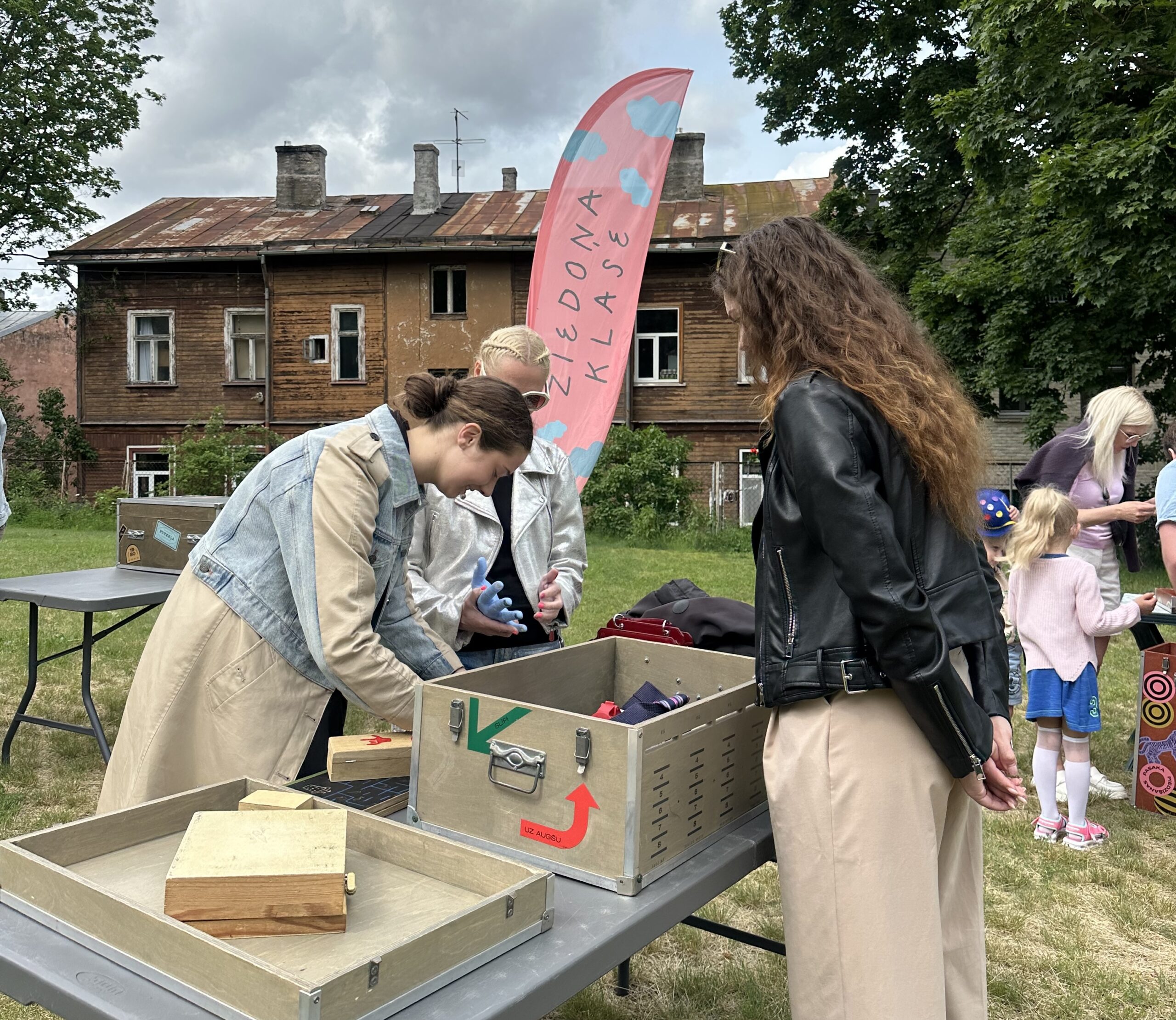 Meitenes stāv ārā pie galda, uz kura ir kastes un dažādi citi priekšmeti. Viņas kaut ko meklē.