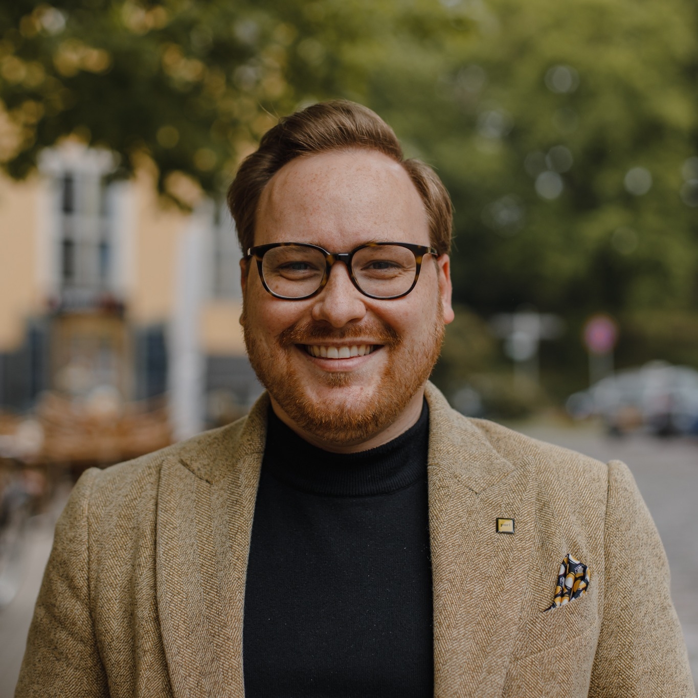 Smaidošs vīrietis Miks Celmiņš ar brillēm un bārdu, ģērbies brūnā žaketē, fonā iela ar kokiem.