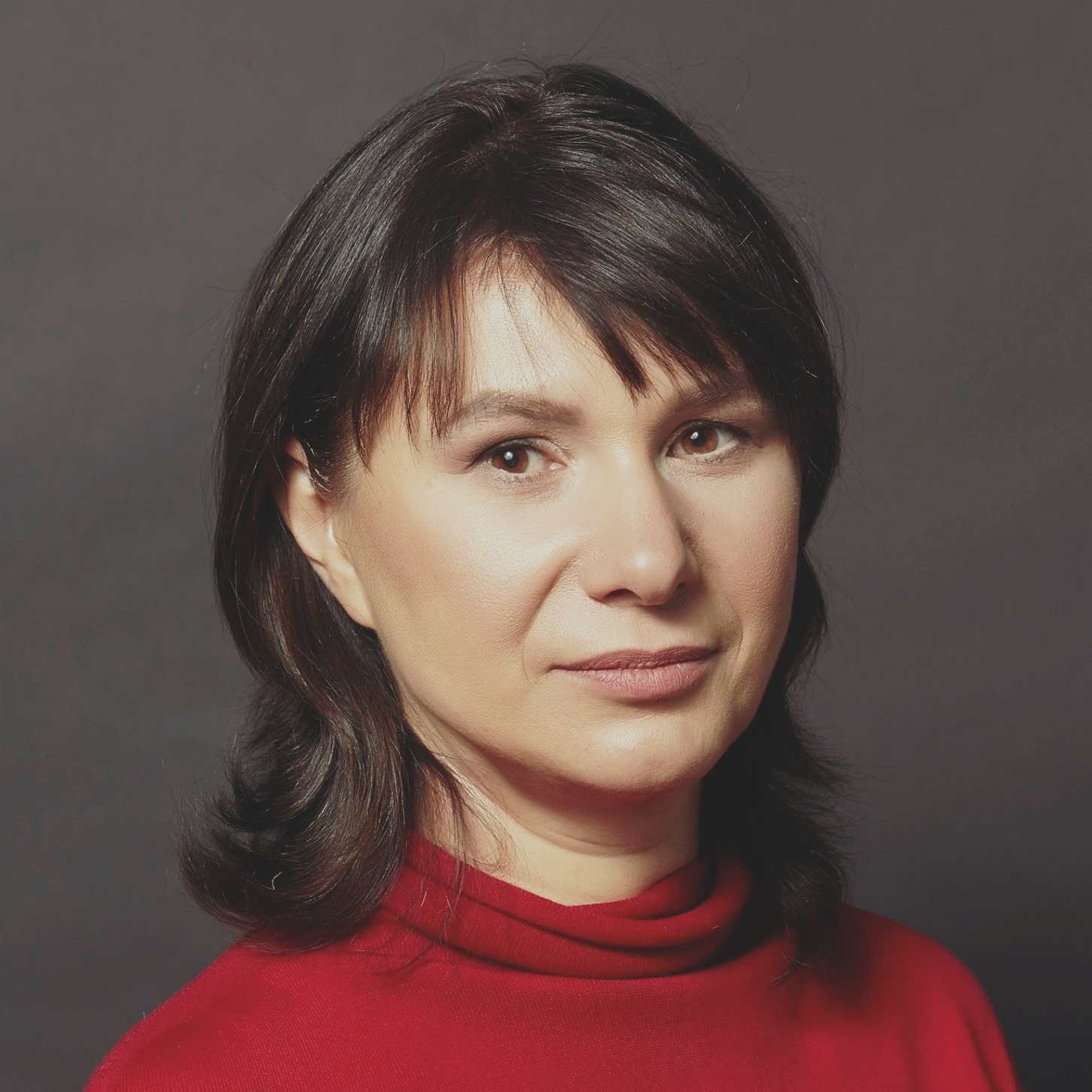 Sieviete Ludmila Kasperoviča ar taisniem tumšiem matiem un sarkanu džemperi skatās kamerā.