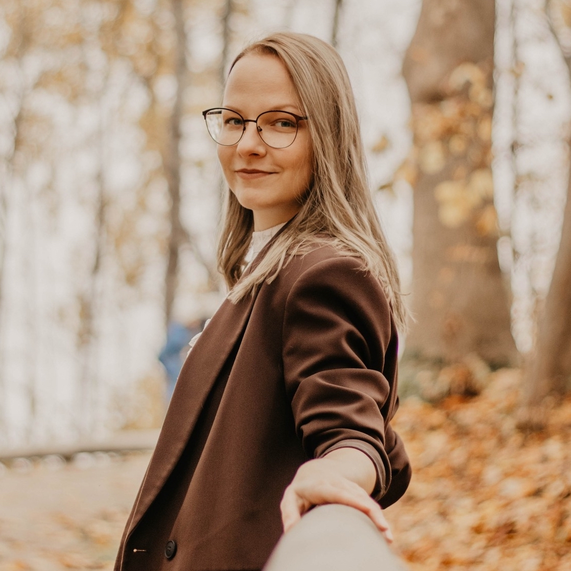 Jauna sieviete Māra Skuja ar brillēm rudens mežā sniedz roku kamerai, smaida.