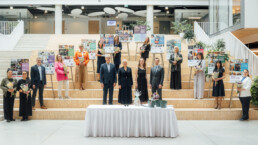 Uz lielām, platām kāpnēm augšup piramīdas formā ir novieteotas 10 planšetes ar fotogrāfijām un pie katras no tām stāv izstādes varoņi - sociālie uzņēmēji. Priekšā viņiem stāv izstādes organizatori - LSUA direktore Regita Zeiļa, pārstāvji no ALTUM, Labklājības ministrijas un Luminor bankas.