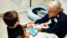 Terapeite sēž pie neliela galdiņa ar mazu zēnu. Abi tur rokās papīra kartiņu. Terapeite tur mazā bērna roku.