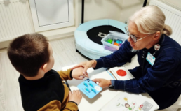 Terapeite sēž pie neliela galdiņa ar mazu zēnu. Abi tur rokās papīra kartiņu. Terapeite tur mazā bērna roku.