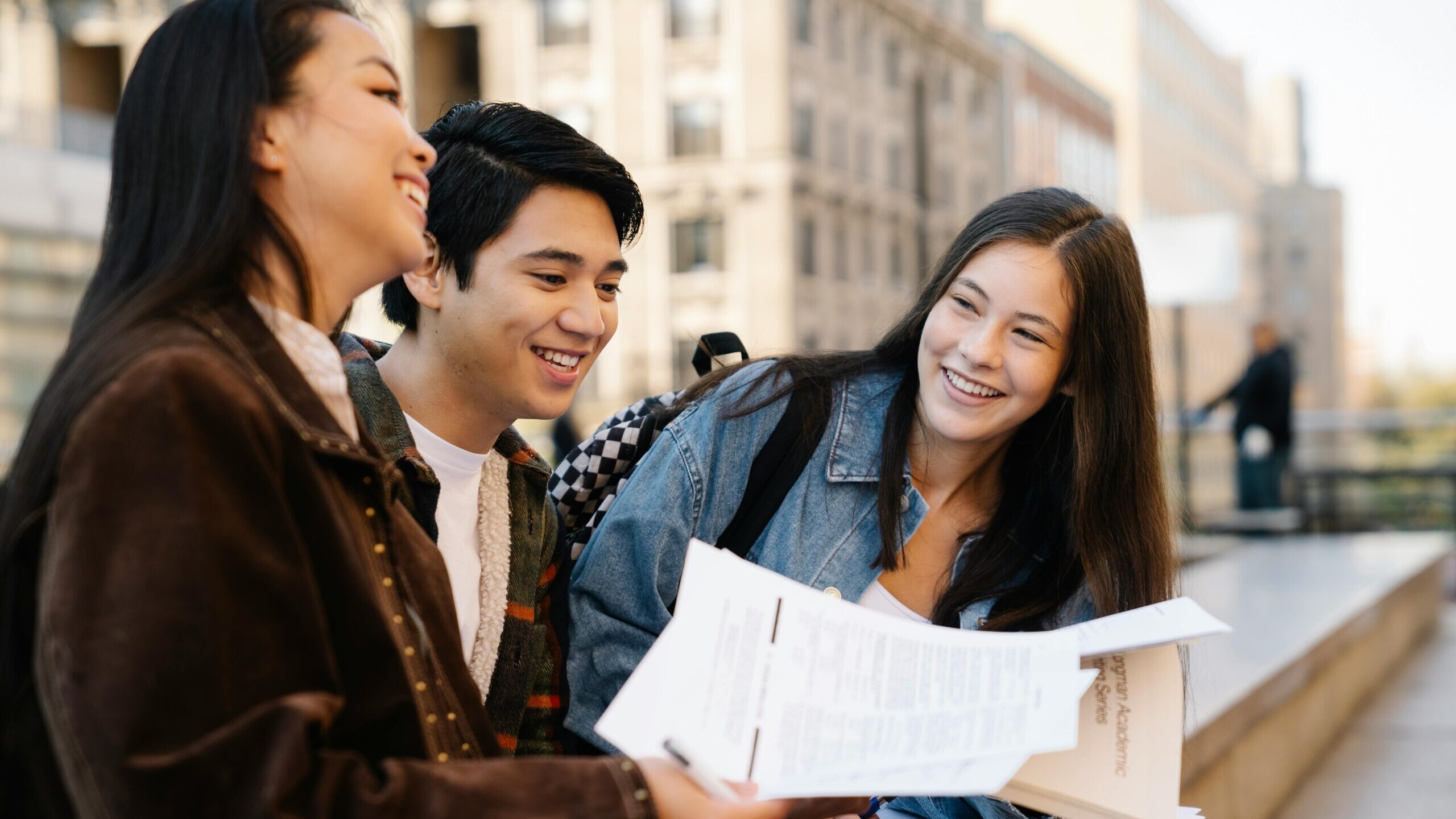 Ilustratīvs attēls. Trīs jaunietes sēž ārā, tur rokā papīra lapas un smaida